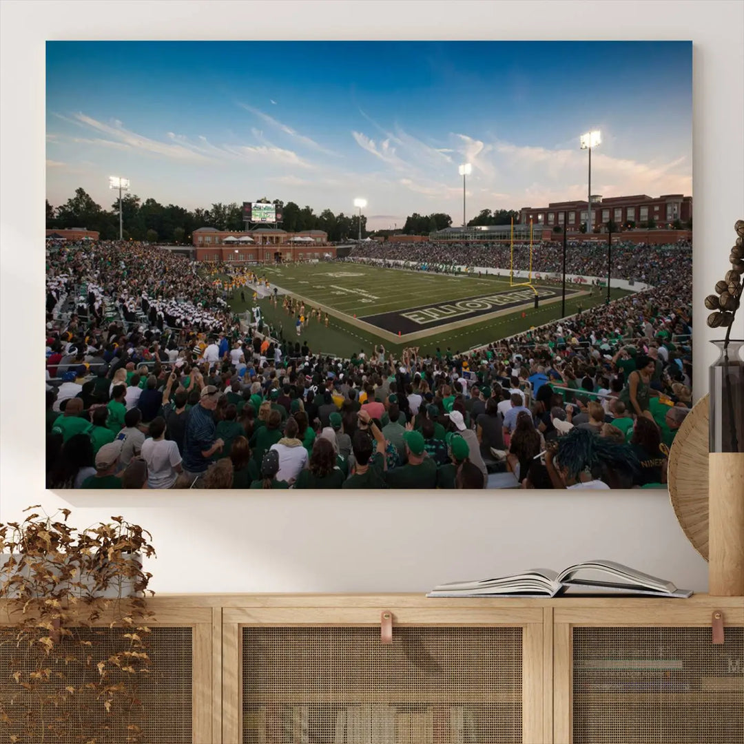 Above the dining table hangs a premium canvas triptych depicting Charlotte Jerry Richardson Stadium, home of the University of Charlotte 49ers Football Team. The scene is alive with a nighttime atmosphere, bustling with a large crowd under bright stadium lights. This stunning art piece is expertly handmade in the USA and adds vibrant energy to your living space.