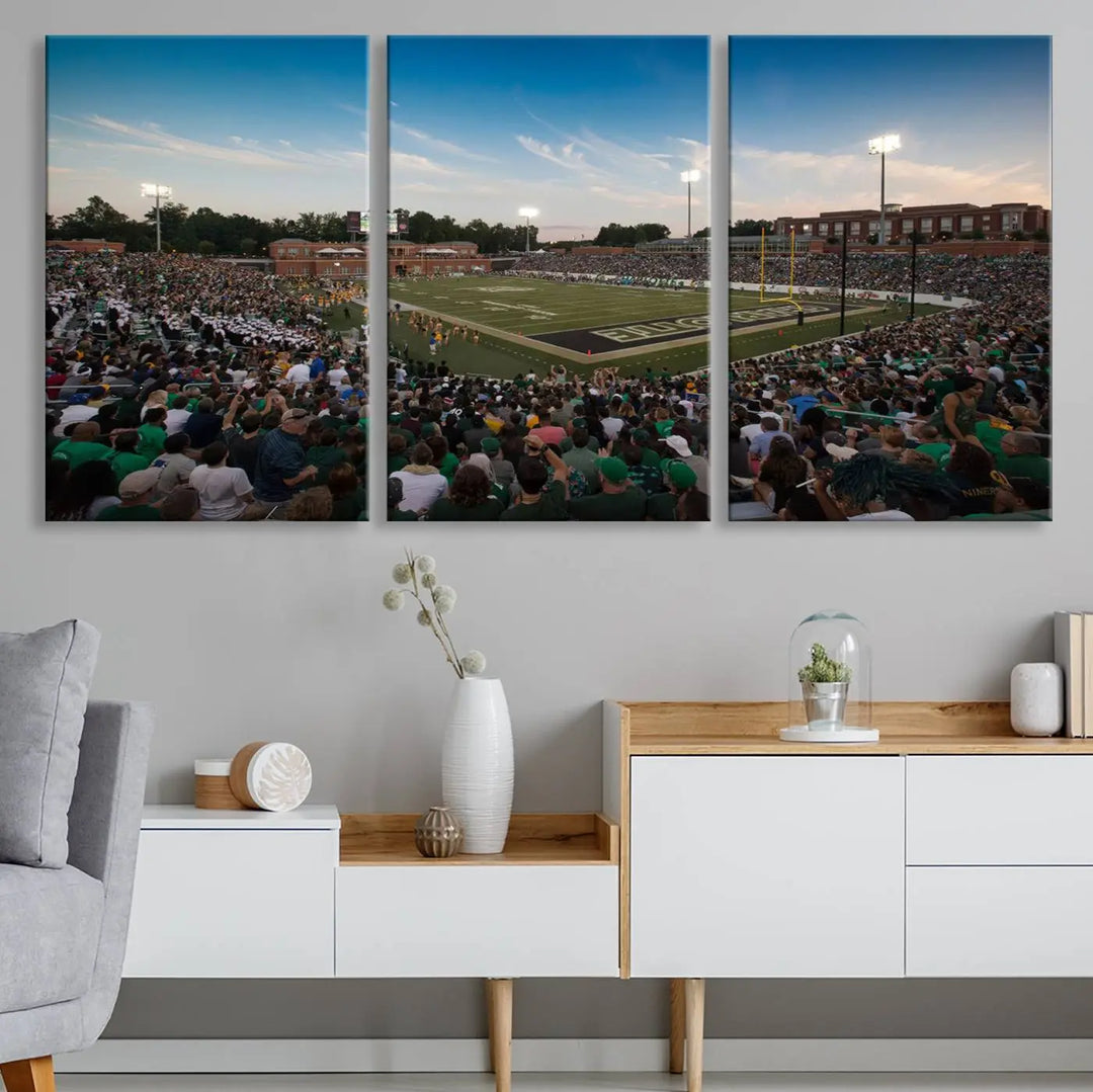 Above the dining table hangs a premium canvas triptych depicting Charlotte Jerry Richardson Stadium, home of the University of Charlotte 49ers Football Team. The scene is alive with a nighttime atmosphere, bustling with a large crowd under bright stadium lights. This stunning art piece is expertly handmade in the USA and adds vibrant energy to your living space.