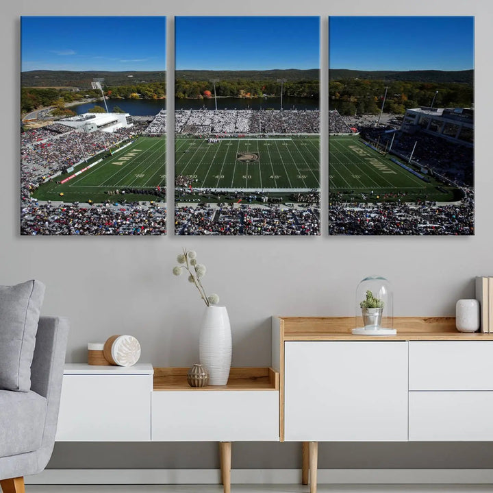 The Army Black Knights Football Team Print on West Point Michie Stadium is showcased with an aerial view capturing a lively football stadium surrounded by lush trees and a river. This wall art is printed on museum-quality canvas and mounted on a stretched wood frame.