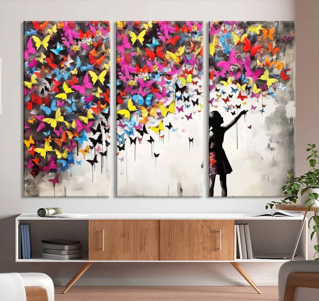 The Banksy Style Wall Art - Girl and Butterfly Explosion features a silhouette of a girl reaching for colorful butterflies on a three-panel canvas print.