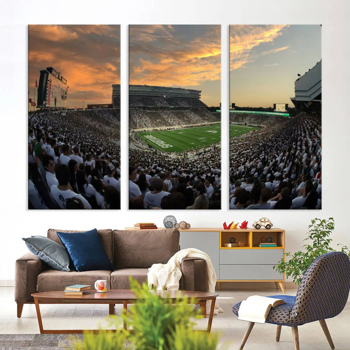 A living room showcasing a stunning Michigan State Spartans Football Team Print on a three-panel canvas, depicting the bustling Spartan Stadium in East Lansing at sunset, all handmade in the USA.