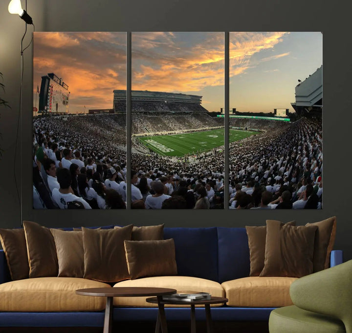 A living room showcasing a stunning Michigan State Spartans Football Team Print on a three-panel canvas, depicting the bustling Spartan Stadium in East Lansing at sunset, all handmade in the USA.