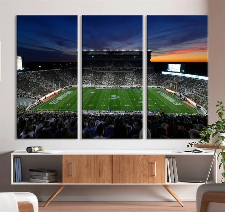 The room showcases a triptych wall art featuring the Michigan State Spartans football team at East Lansing Spartan Stadium at dusk, elegantly printed on premium canvas for a gallery-quality finish.