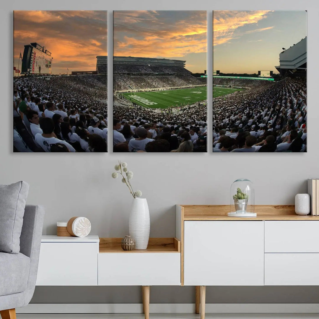 A living room showcasing a stunning Michigan State Spartans Football Team Print on a three-panel canvas, depicting the bustling Spartan Stadium in East Lansing at sunset, all handmade in the USA.