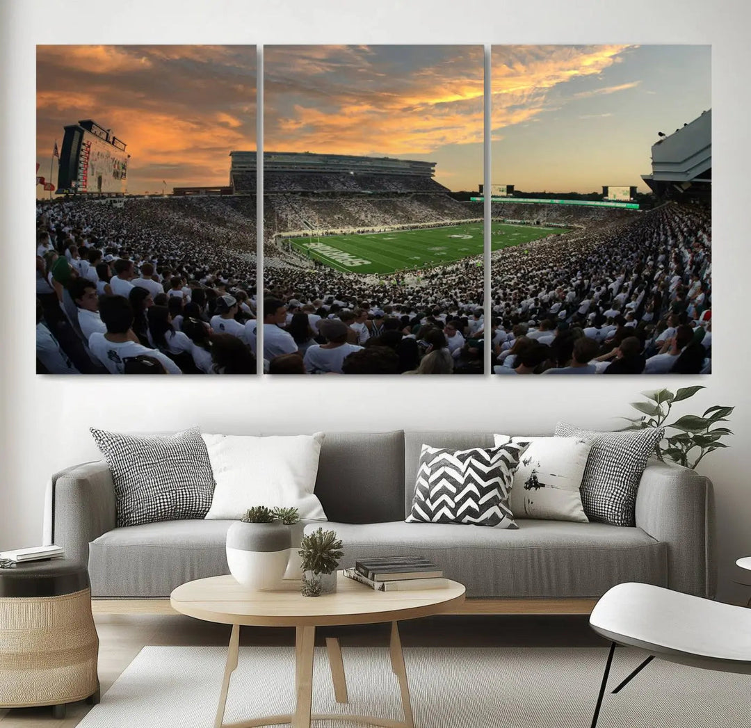 A living room showcasing a stunning Michigan State Spartans Football Team Print on a three-panel canvas, depicting the bustling Spartan Stadium in East Lansing at sunset, all handmade in the USA.