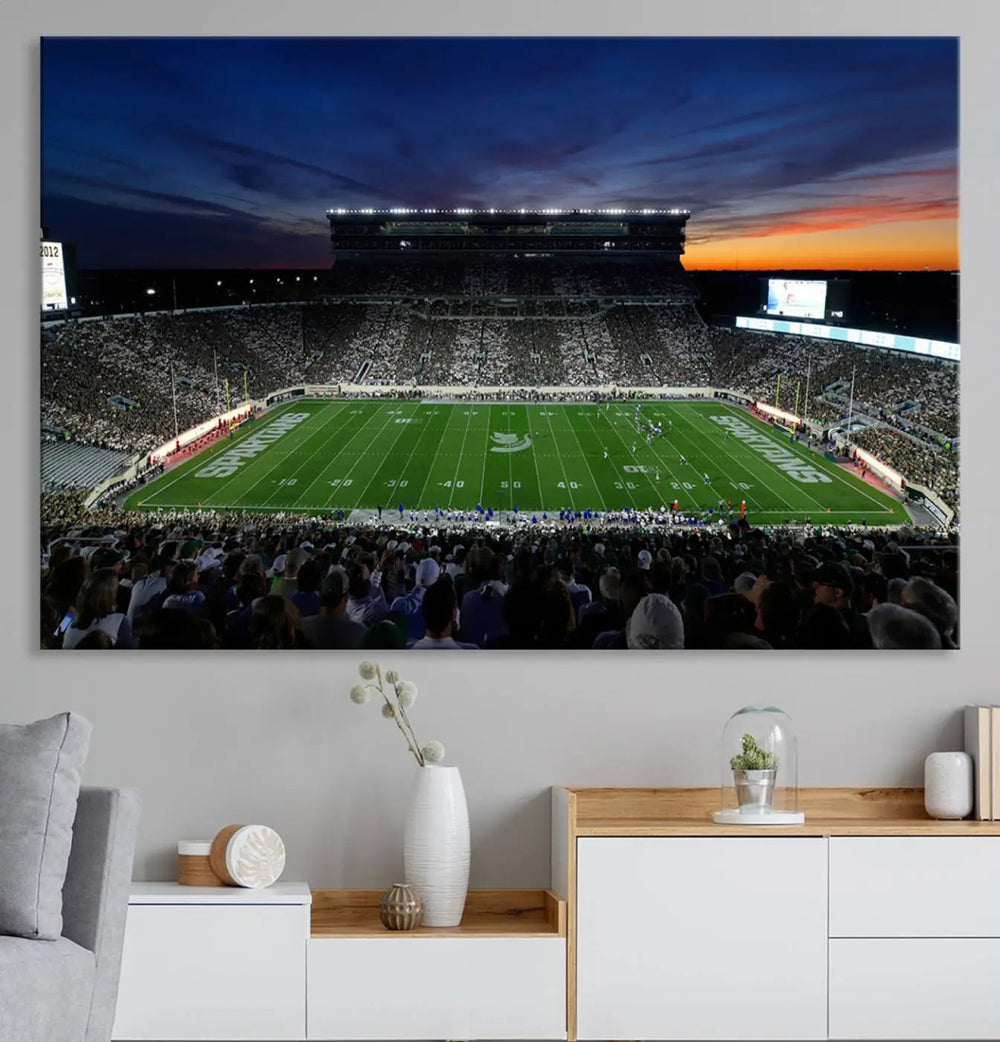 The room showcases a triptych wall art featuring the Michigan State Spartans football team at East Lansing Spartan Stadium at dusk, elegantly printed on premium canvas for a gallery-quality finish.