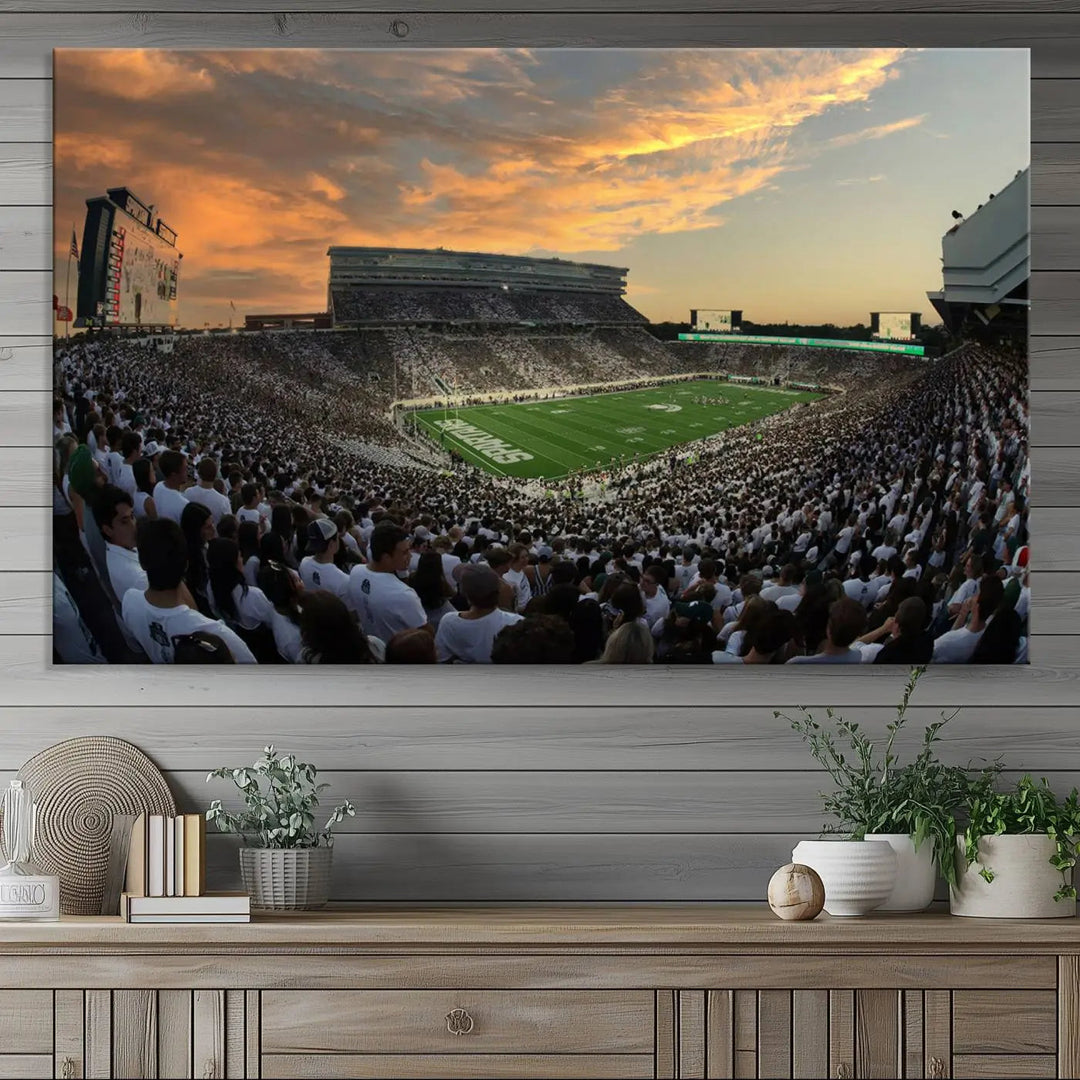 A living room showcasing a stunning Michigan State Spartans Football Team Print on a three-panel canvas, depicting the bustling Spartan Stadium in East Lansing at sunset, all handmade in the USA.