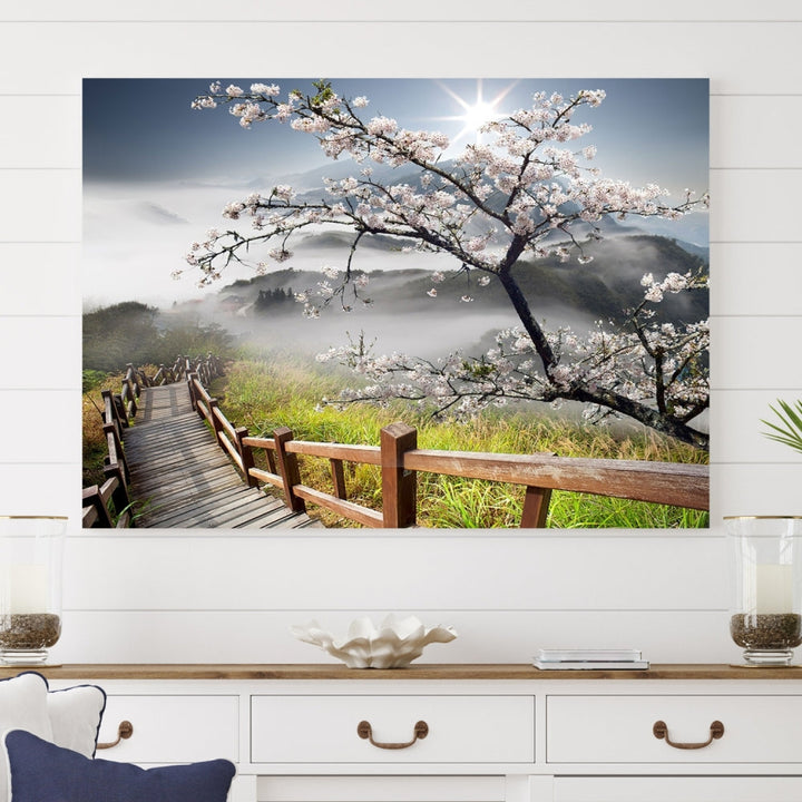 The wall art depicts a sunlit cherry blossom tree forming a tranquil canopy over a wooden path winding through misty hills. This serene image captures the essence of a walk through blossoming nature, reminiscent of Sakura Cherry Blossom Pathway Canvas Wall Art, and is ready to hang.