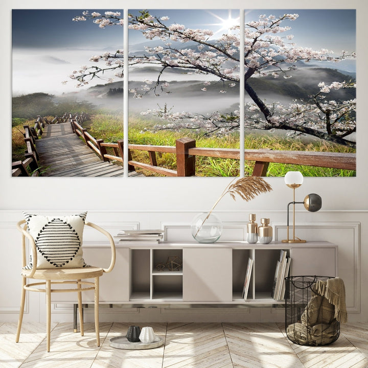 The wall art depicts a sunlit cherry blossom tree forming a tranquil canopy over a wooden path winding through misty hills. This serene image captures the essence of a walk through blossoming nature, reminiscent of Sakura Cherry Blossom Pathway Canvas Wall Art, and is ready to hang.