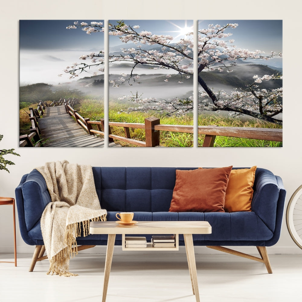 The wall art depicts a sunlit cherry blossom tree forming a tranquil canopy over a wooden path winding through misty hills. This serene image captures the essence of a walk through blossoming nature, reminiscent of Sakura Cherry Blossom Pathway Canvas Wall Art, and is ready to hang.