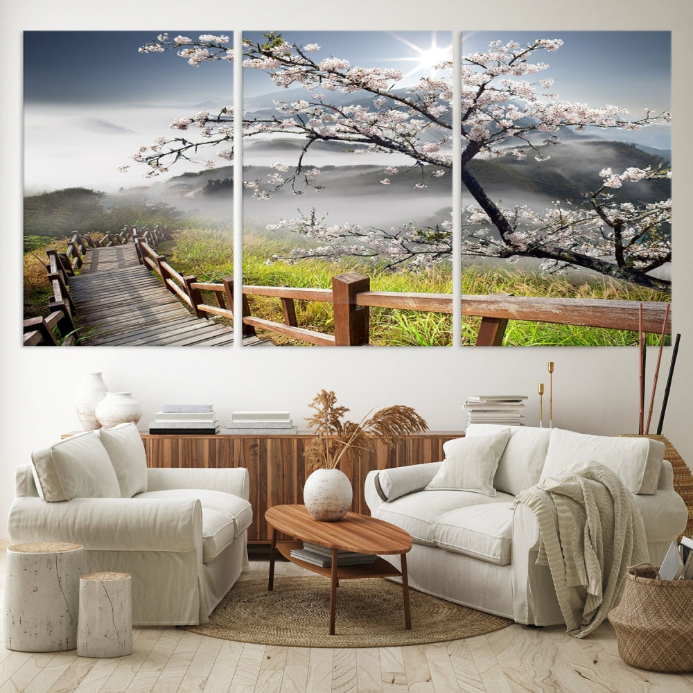 The wall art depicts a sunlit cherry blossom tree forming a tranquil canopy over a wooden path winding through misty hills. This serene image captures the essence of a walk through blossoming nature, reminiscent of Sakura Cherry Blossom Pathway Canvas Wall Art, and is ready to hang.