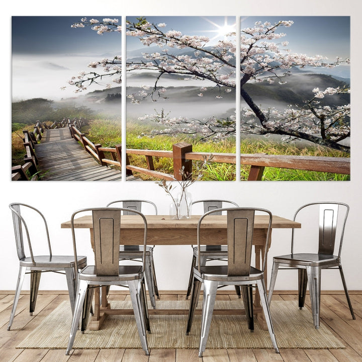 The wall art depicts a sunlit cherry blossom tree forming a tranquil canopy over a wooden path winding through misty hills. This serene image captures the essence of a walk through blossoming nature, reminiscent of Sakura Cherry Blossom Pathway Canvas Wall Art, and is ready to hang.