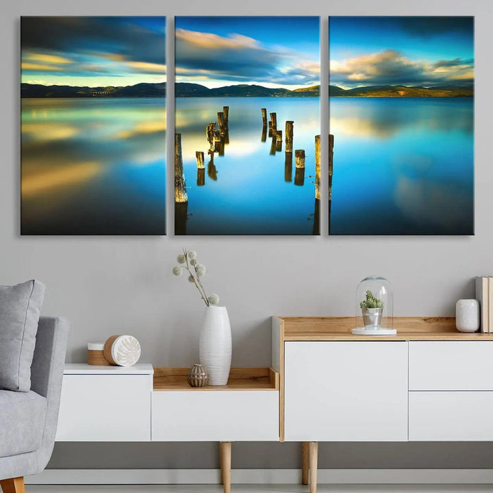 A triptych of a serene lake scene with old wooden pillars, blue sky, and clouds casts a coastal ambiance. The Sea Ocean Sunset Beach Wall Art Canvas Print reflects the soothing nature of the ocean.