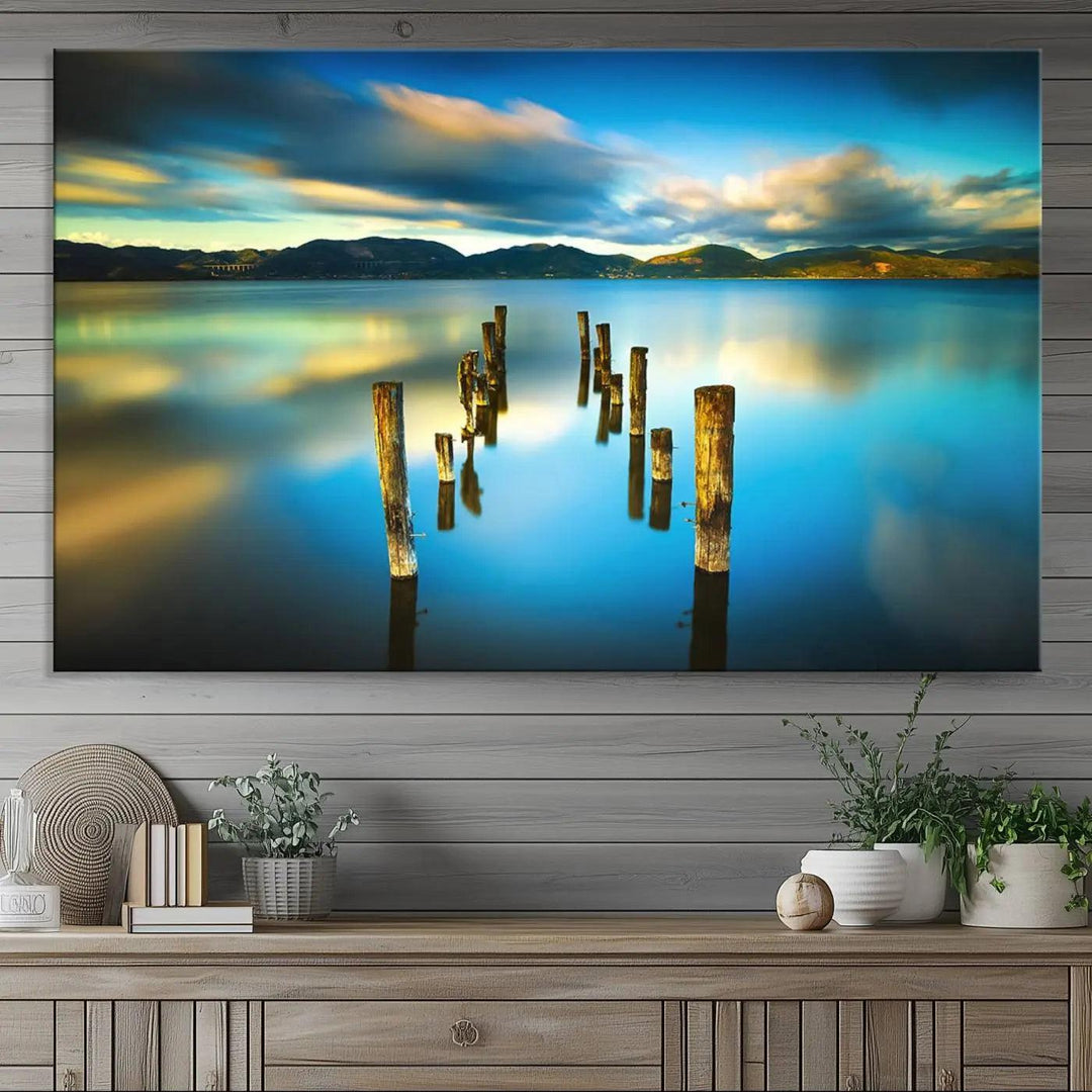 A triptych of a serene lake scene with old wooden pillars, blue sky, and clouds casts a coastal ambiance. The Sea Ocean Sunset Beach Wall Art Canvas Print reflects the soothing nature of the ocean.