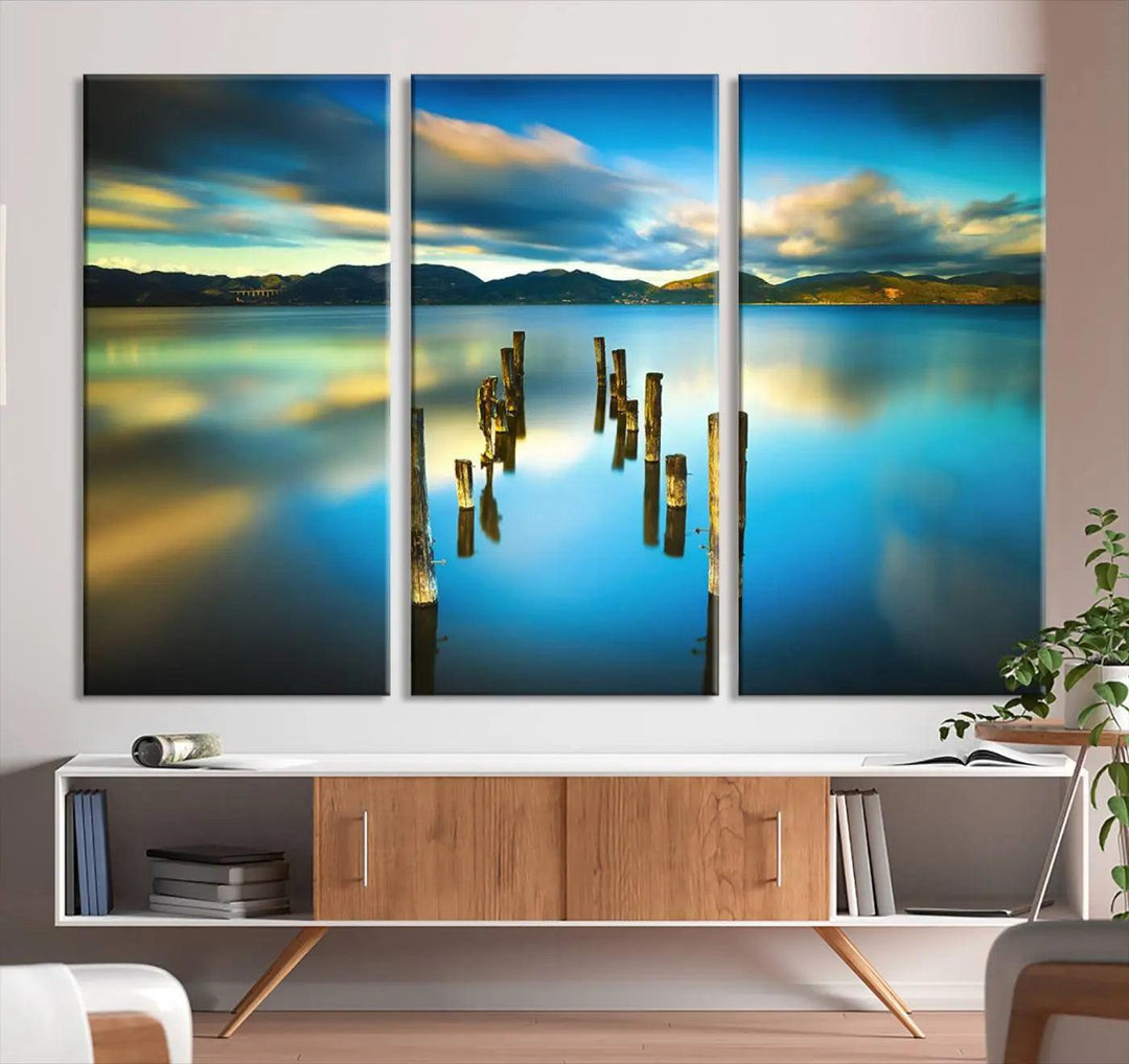 A triptych of a serene lake scene with old wooden pillars, blue sky, and clouds casts a coastal ambiance. The Sea Ocean Sunset Beach Wall Art Canvas Print reflects the soothing nature of the ocean.