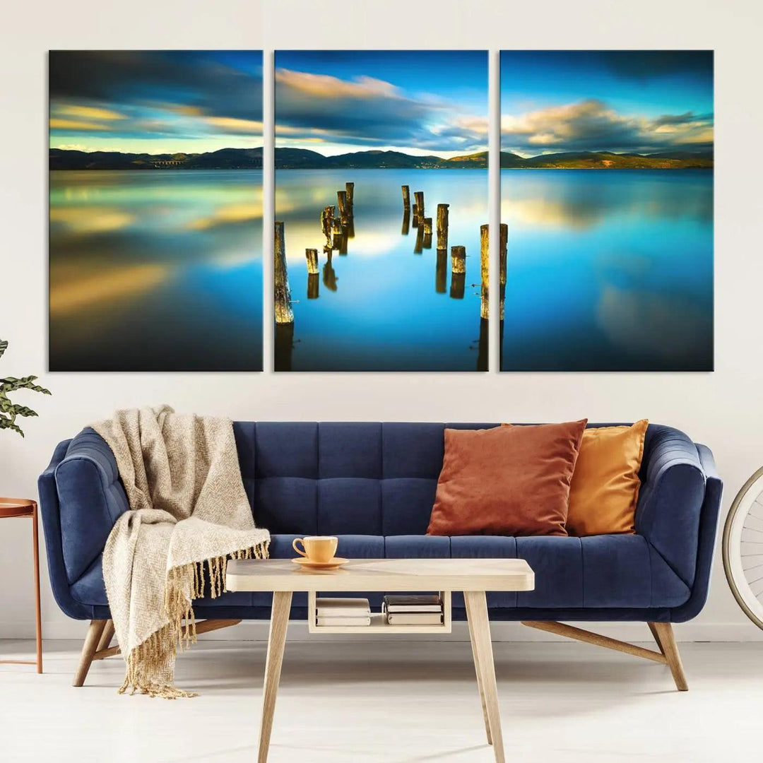 A triptych of a serene lake scene with old wooden pillars, blue sky, and clouds casts a coastal ambiance. The Sea Ocean Sunset Beach Wall Art Canvas Print reflects the soothing nature of the ocean.