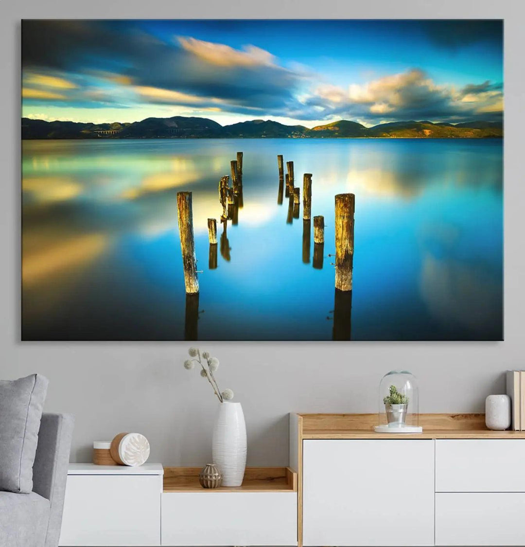 A triptych of a serene lake scene with old wooden pillars, blue sky, and clouds casts a coastal ambiance. The Sea Ocean Sunset Beach Wall Art Canvas Print reflects the soothing nature of the ocean.