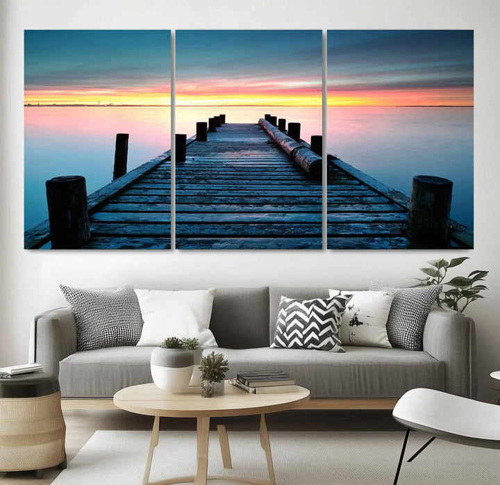 The "Sunset Pier Triptych Canvas Art," a giclee print known for its Canon print quality and gallery wrap finish, captures a wooden pier stretching into a tranquil lake at sunset beneath vibrant orange and blue skies. It makes an ideal centerpiece for coastal-themed art settings.