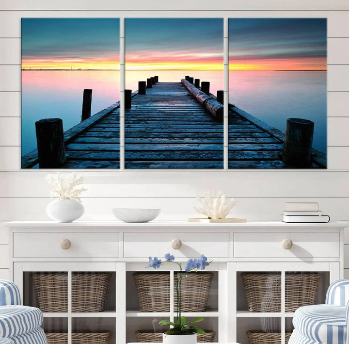 The "Sunset Pier Triptych Canvas Art," a giclee print known for its Canon print quality and gallery wrap finish, captures a wooden pier stretching into a tranquil lake at sunset beneath vibrant orange and blue skies. It makes an ideal centerpiece for coastal-themed art settings.