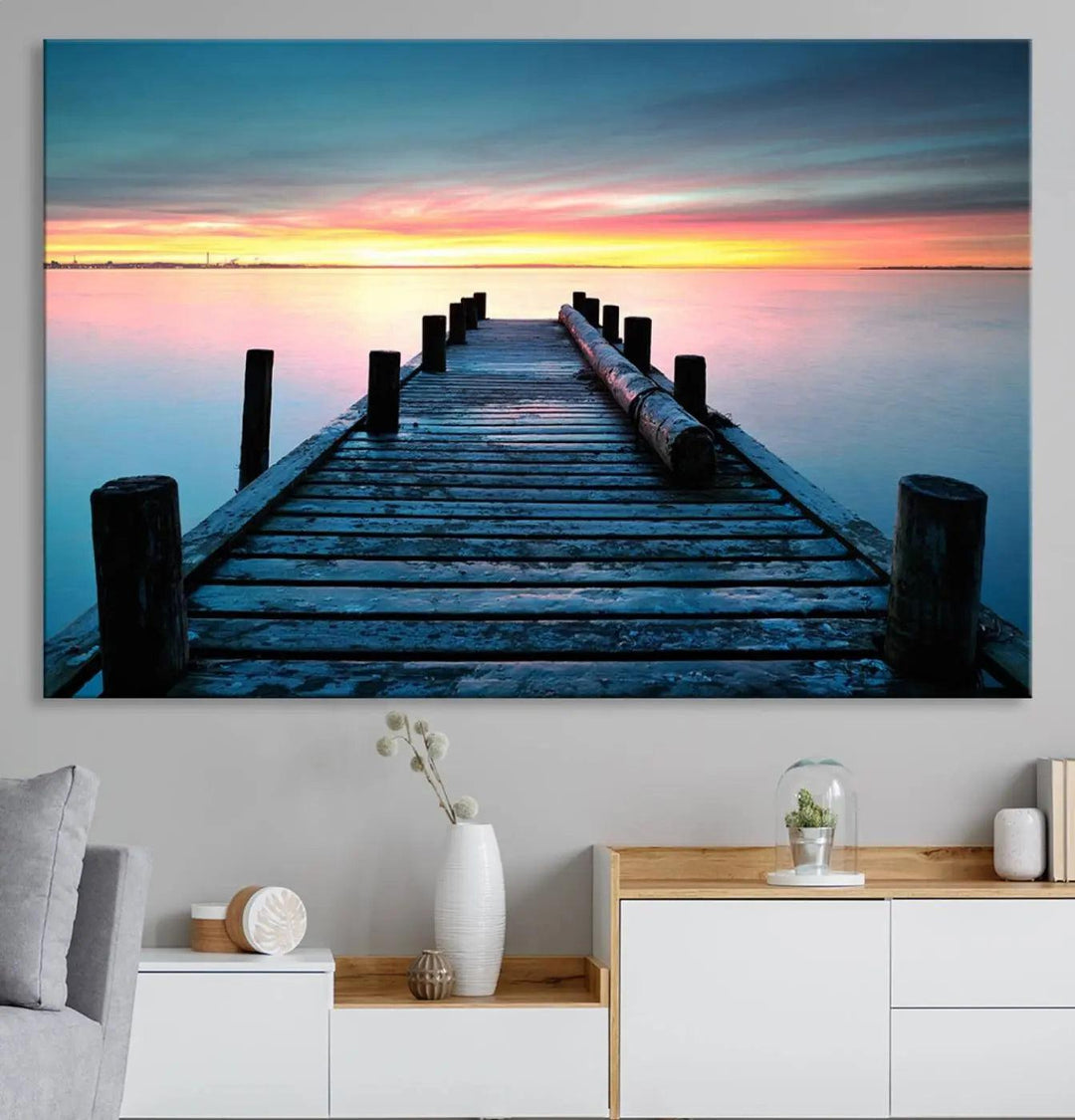 The "Sunset Pier Triptych Canvas Art," a giclee print known for its Canon print quality and gallery wrap finish, captures a wooden pier stretching into a tranquil lake at sunset beneath vibrant orange and blue skies. It makes an ideal centerpiece for coastal-themed art settings.