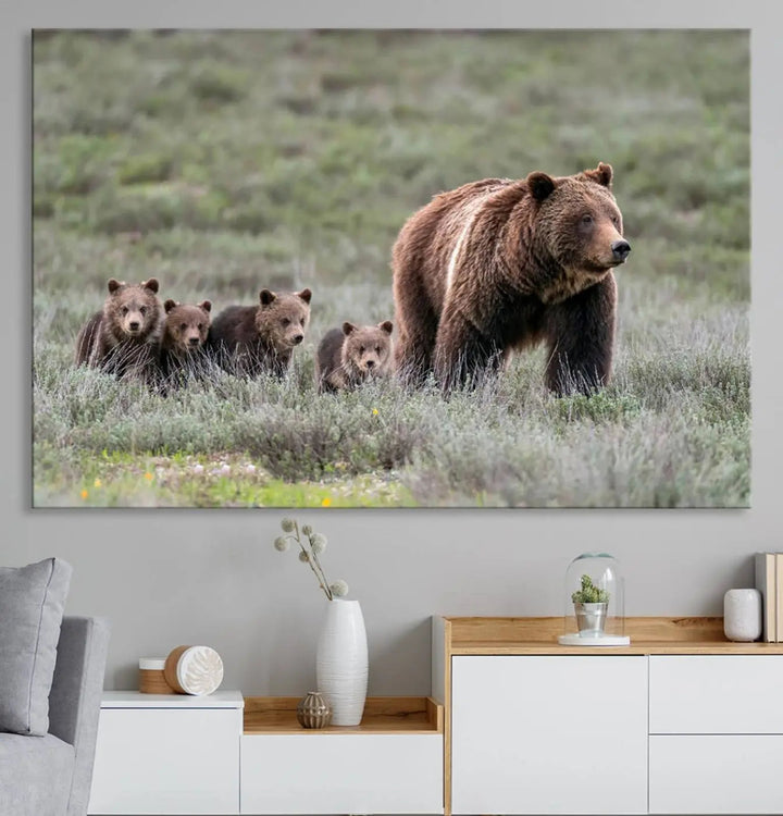 The canvas wall art print, titled "Queen of the Tetons," features a photo of 399 grizzly bear cubs walking with a larger bear in a green, open field. This striking piece is framed and leaning against a wall, showcasing its gallery-quality finish.